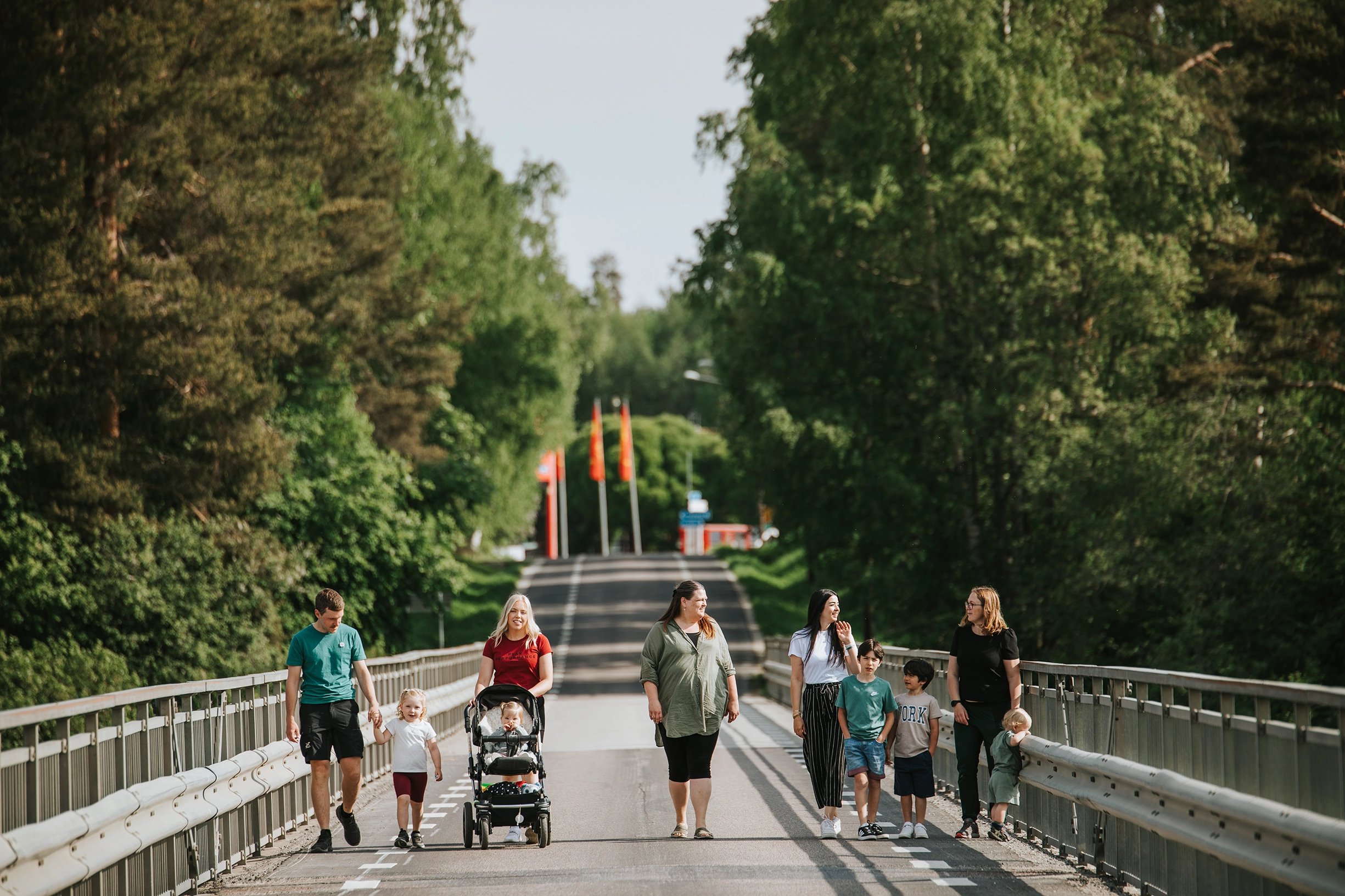 Barn och vuxna gående eller åkandes i vagn på en bro omgiven av grönska.