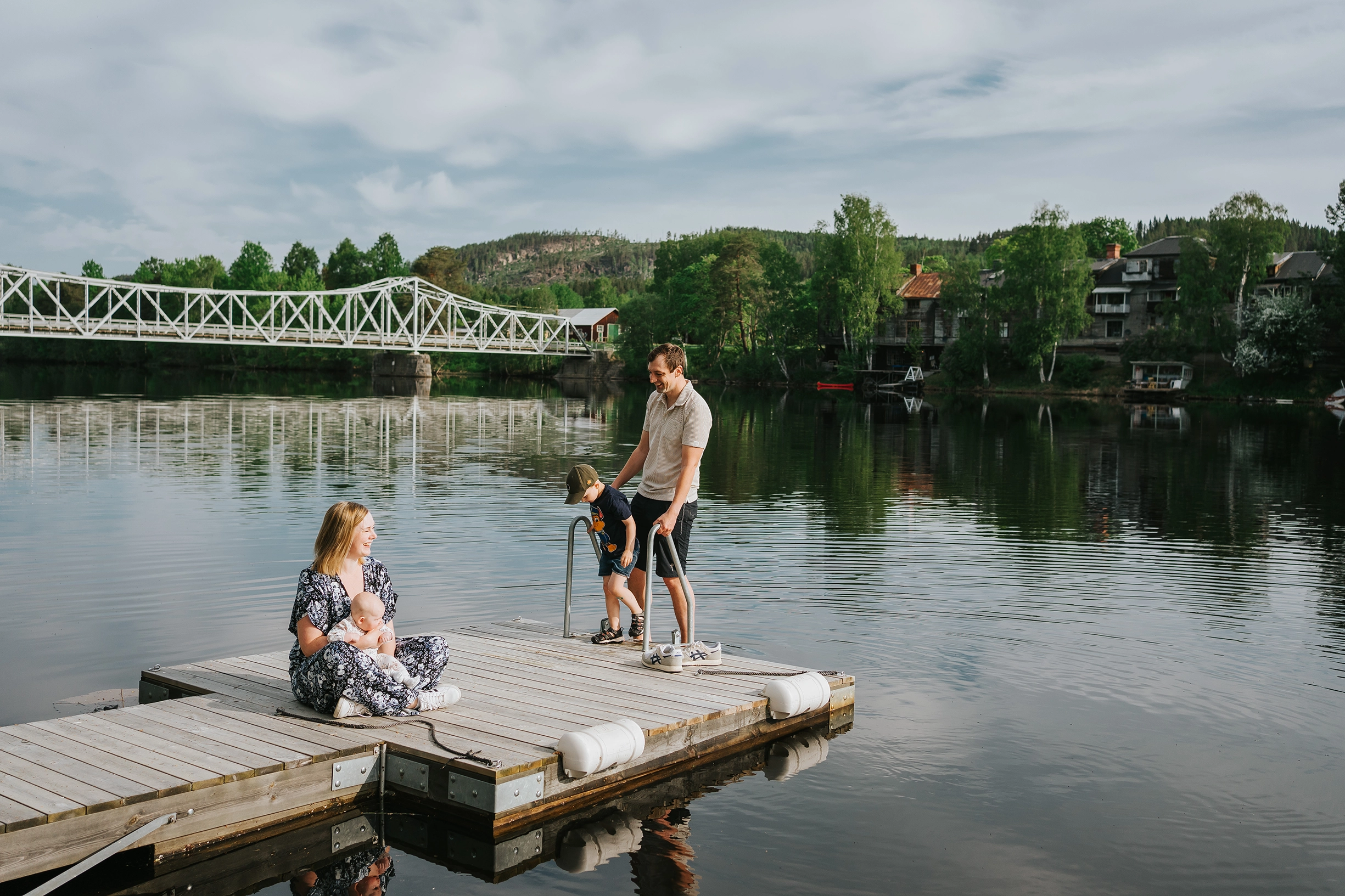 Två vuxna och ett barn på en brygga omgiven av vatten leendes mot varandra. I bakgrunden syns en vit bro.