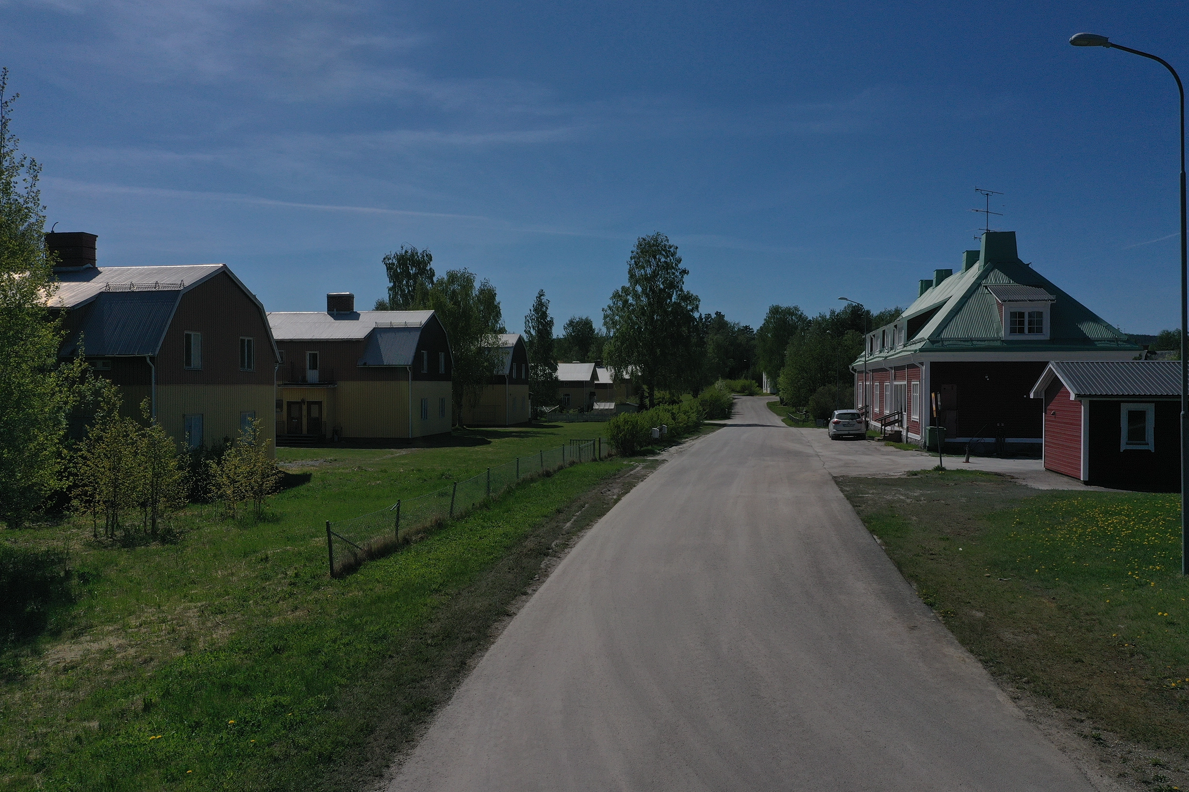 En grusväg. På höger sida är ett rött hus med vita knutar. På andra sidan vägen står tre stycken likadana gula och bruna villor.