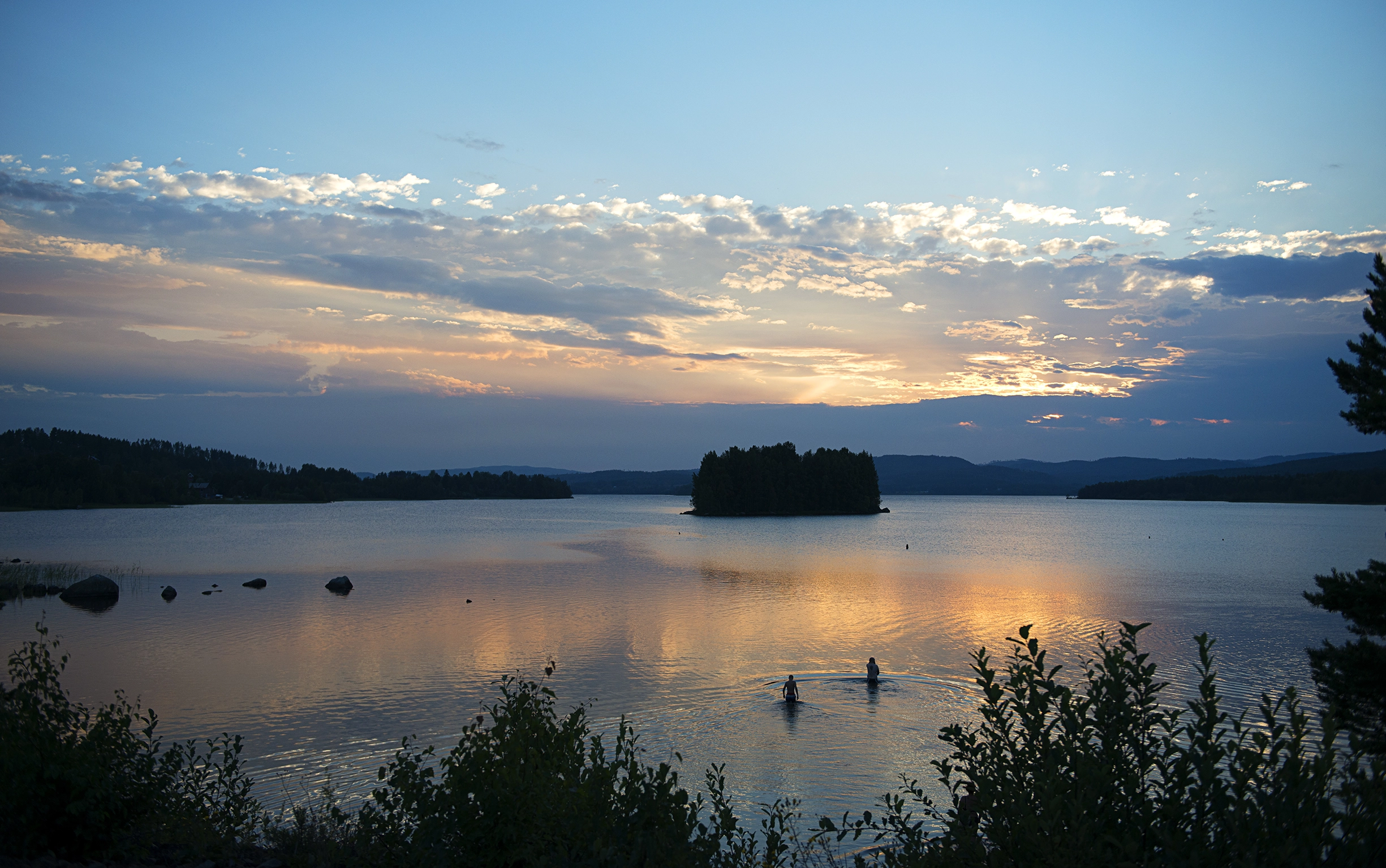 Två stycken personer badar i en sjö omgiven av grönska i solnedgången