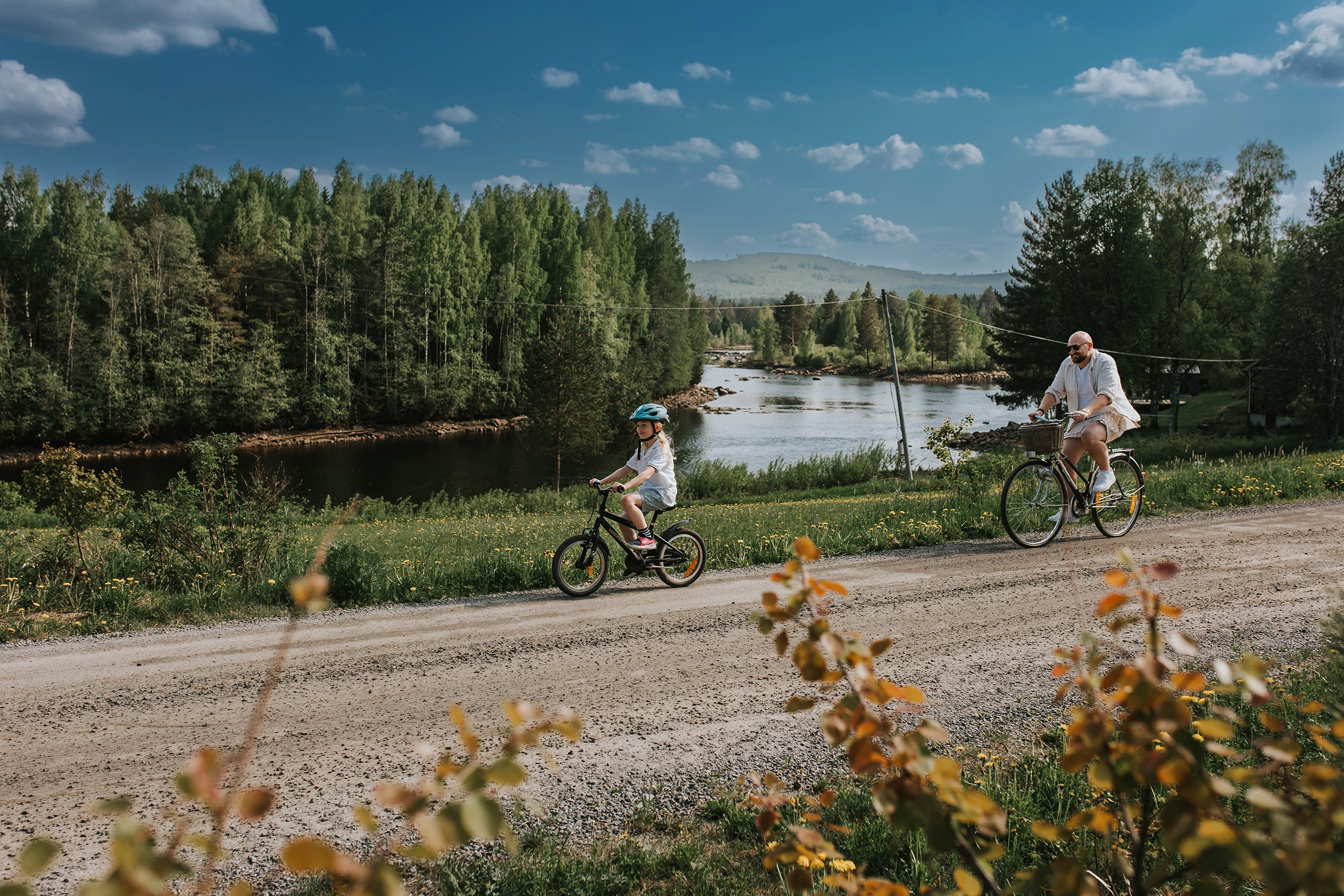 En vuxen och ett barn cyklar på varsin cykel på en grusväg omgiven av grönska och bredvid forsar vatten.
