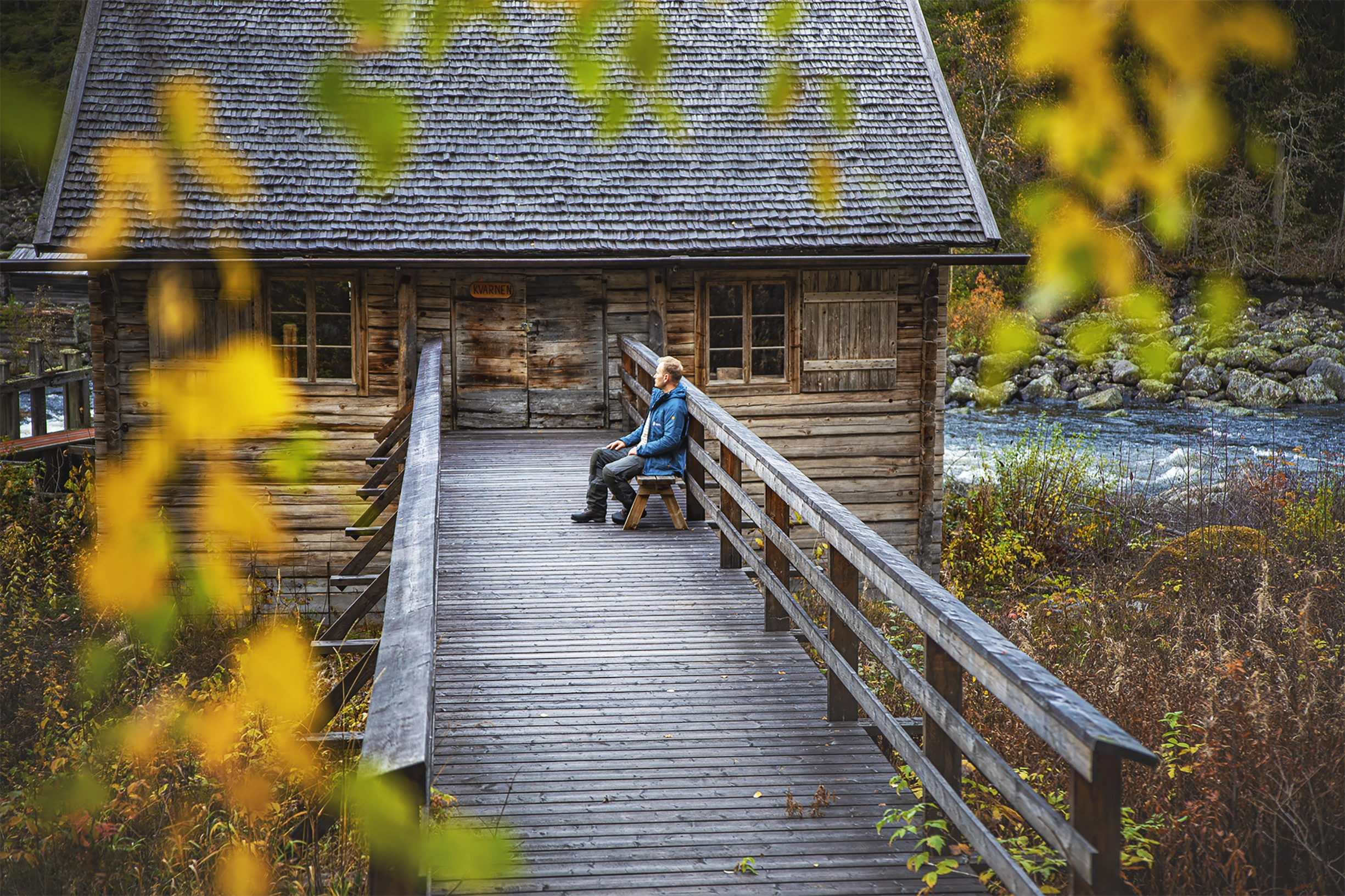 En person sitter på en bänk på en träbrygga som leder fram till ett trähus